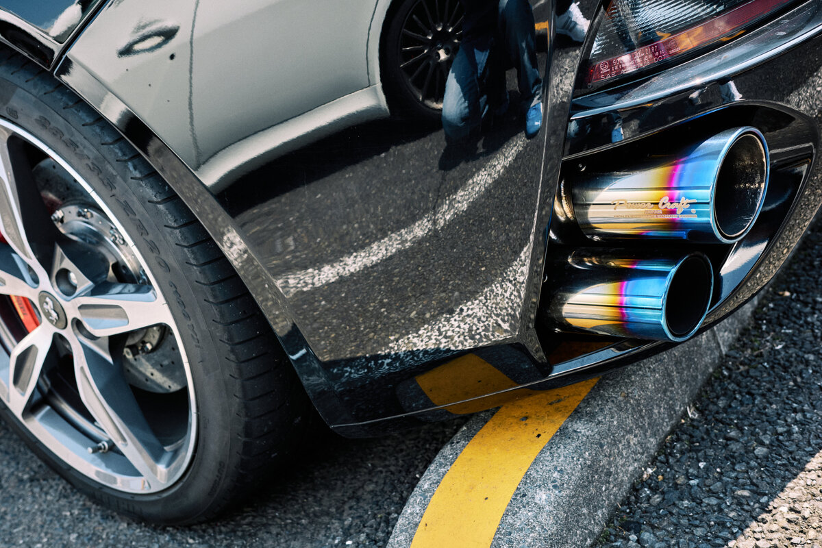 Close-up of a Ferrari's rear wheel and quad burnt titanium exhaust pipes, highlighting the reflective black paint and performance detailing.
