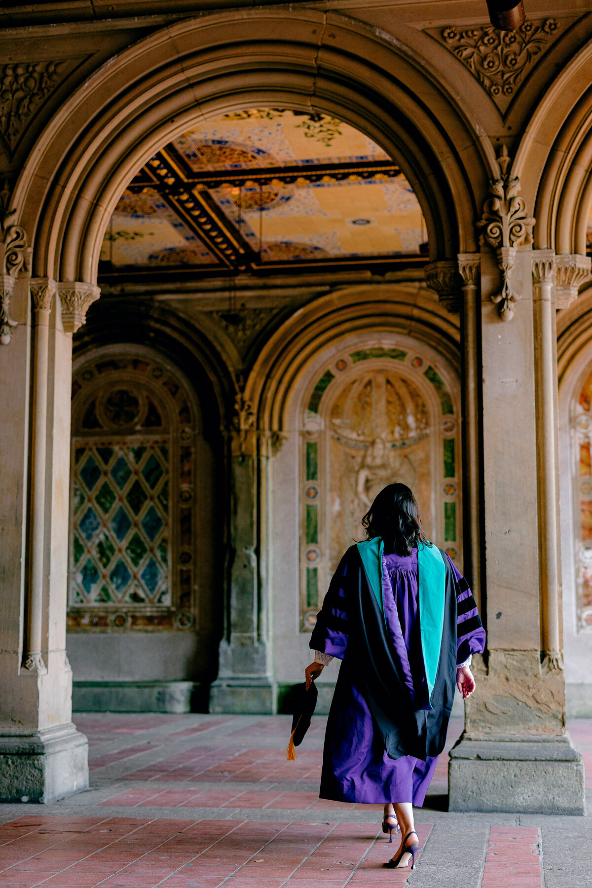 NYU graduate walking through the arches in purple gown and hood at Central Park.