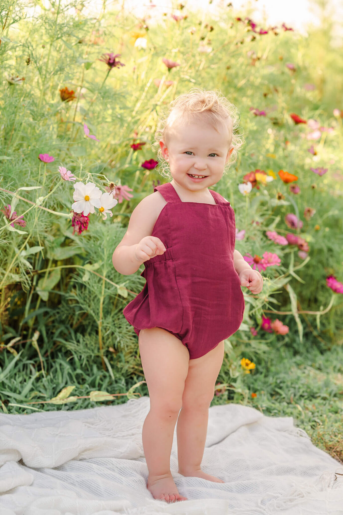 A toddler wearing a red romper smiles for the camera while standing on a blanket in a wildflower field near Moyock, NC.