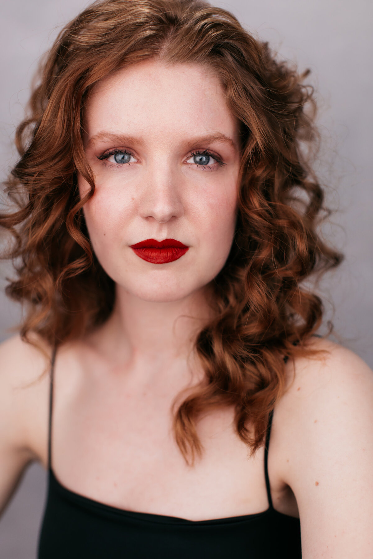 Castingfoto van Anne Homburg. Vrouw met roodbruine krullen en rode lippenstift voor grijze achtergrond.