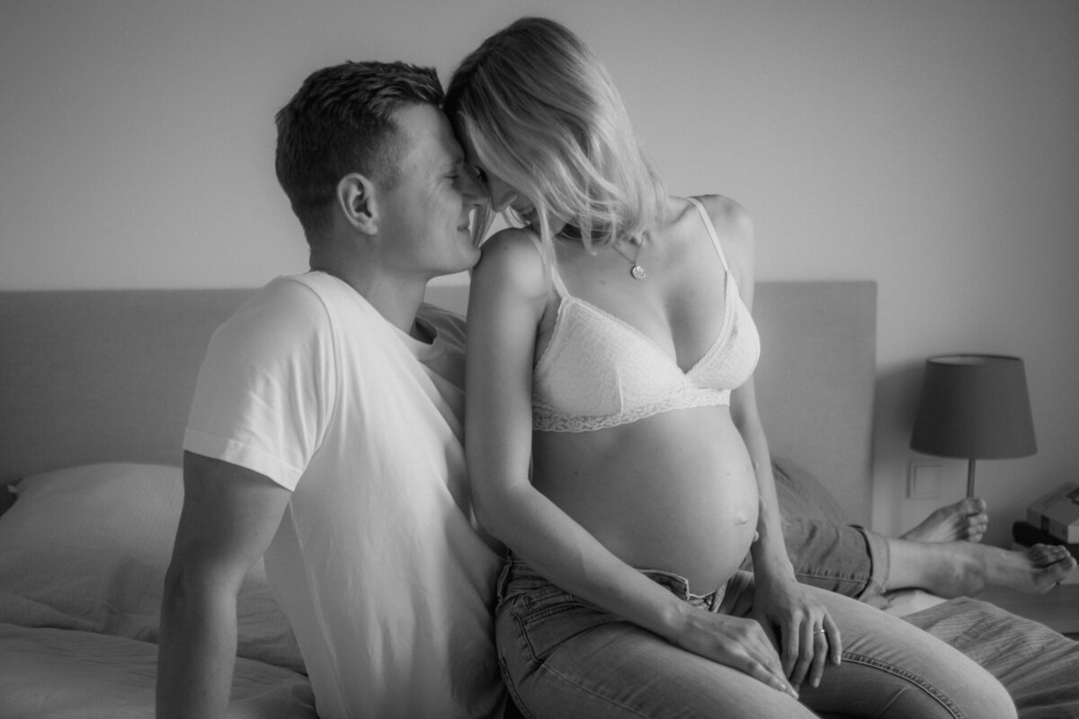 Expecting couple sits closely on a bed.