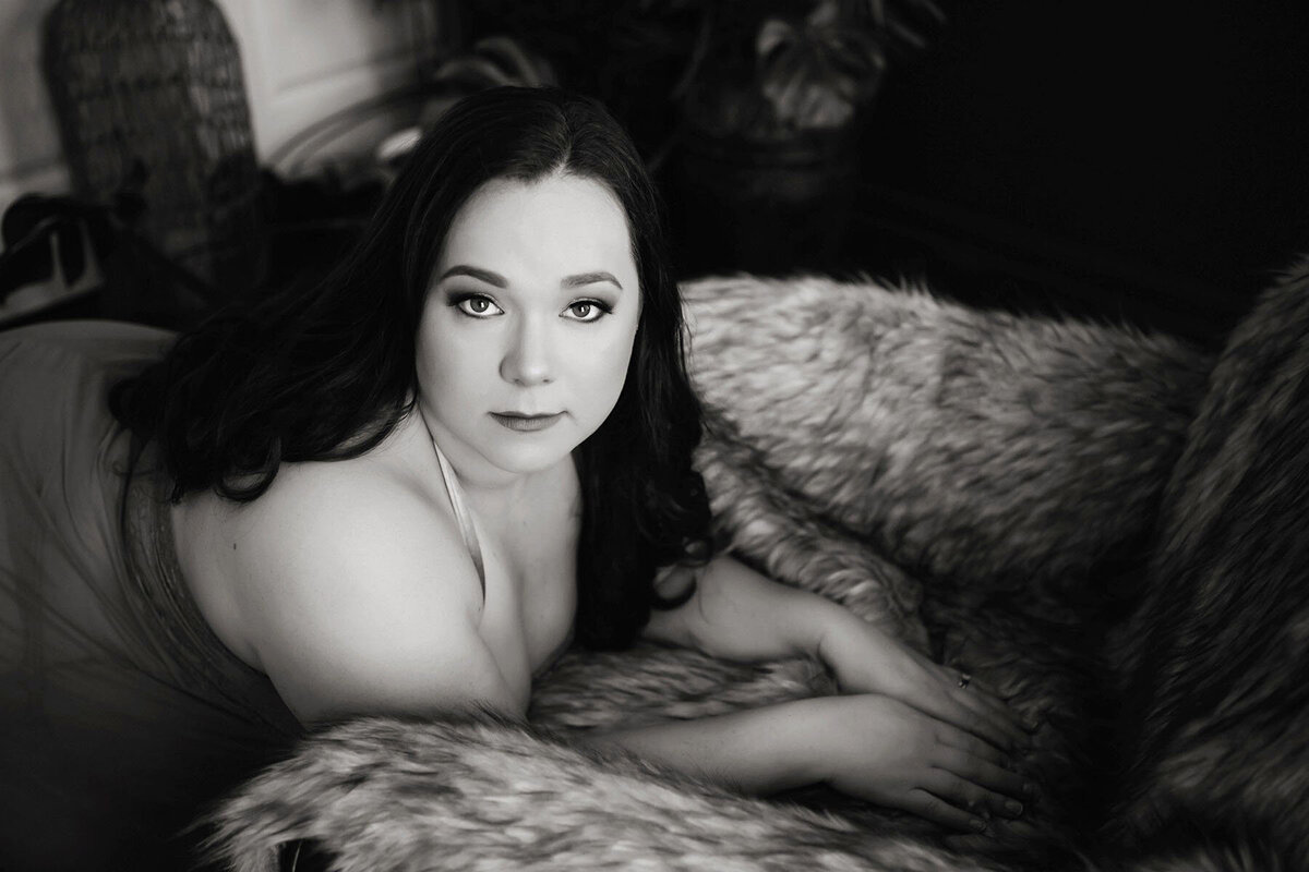 Black and white image of a woman with long dark hair, lying on a furry blanket, looking at the camera with a calm expression. Cozy and serene atmosphere.