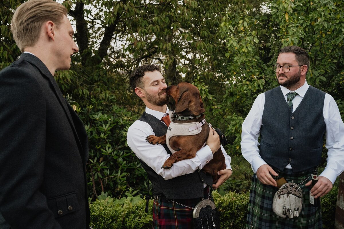 A man holds a sausage dog and the dog licks his face. Two wedding guests stand at either side.