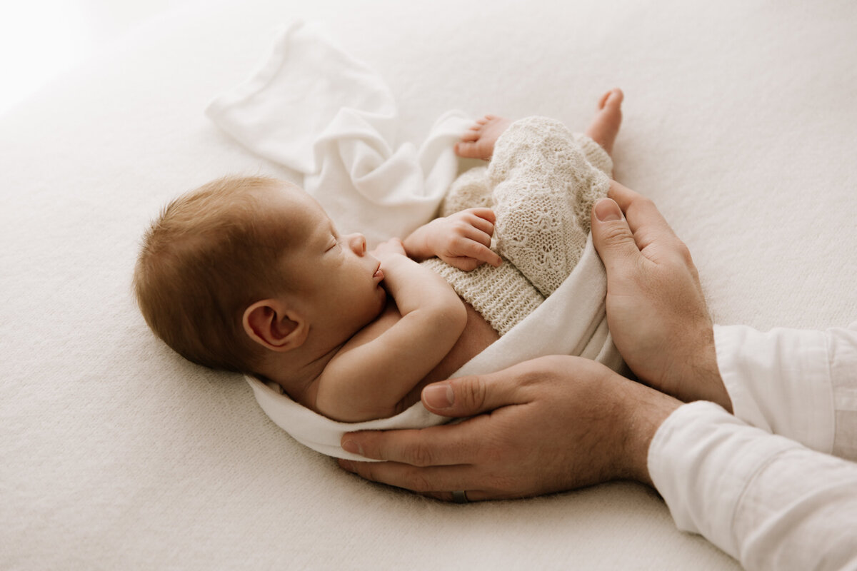 A peaceful newborn baby sleeps on a soft cream blanket, wrapped loosely in a white swaddle and dressed in knitted pants. The baby's tiny body is gently cradled by their father's protective hands, creating a tender and intimate moment. Soft, diffused light highlights the baby's serene features and delicate details. Captured in a Sydney newborn photography studio.