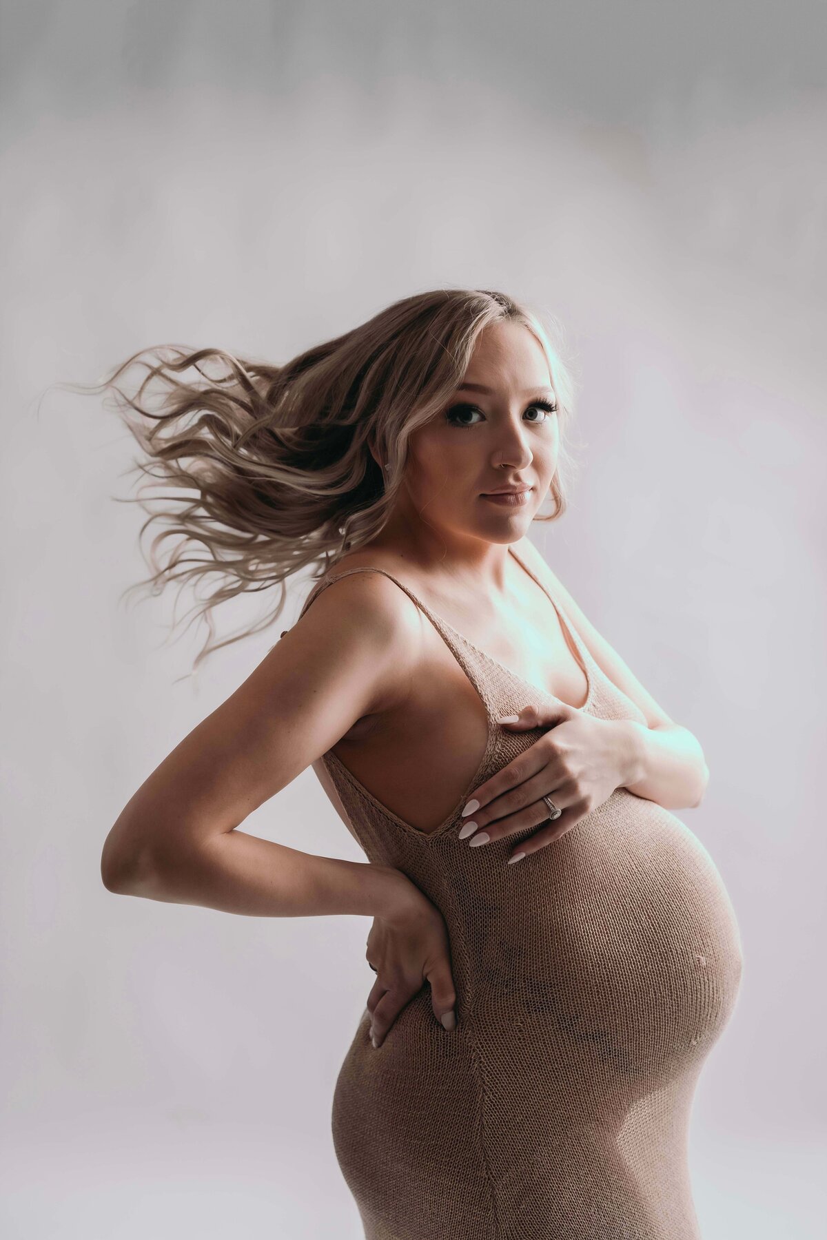 Close-up maternity portrait of an expecting mother in the studio of a Mobile, AL photographer. The image focuses on her upper body as she wears a sheer, modern gown. One hand rests modestly across her chest while the other supports her lower back, highlighting her pregnancy glow. Her hair is flowing behind her for an extra artistic flare.