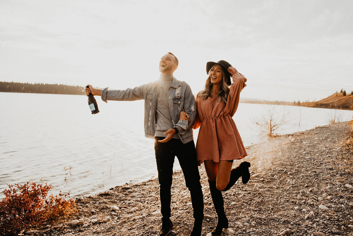 Trendy engaged couple, popping champagne along the beach, captured by Kelsey Vera Photography, intimate and romantic wedding photographer in Airdrie, Alberta. Featured on the Bronte Bride Vendor Guide.