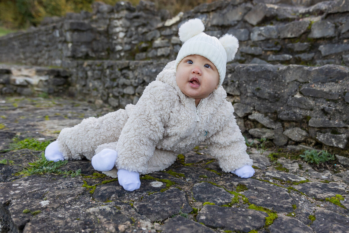 A baby in a white fluffy outfit and hat crawls on a mossy stone surface with ancient ruins in the background.