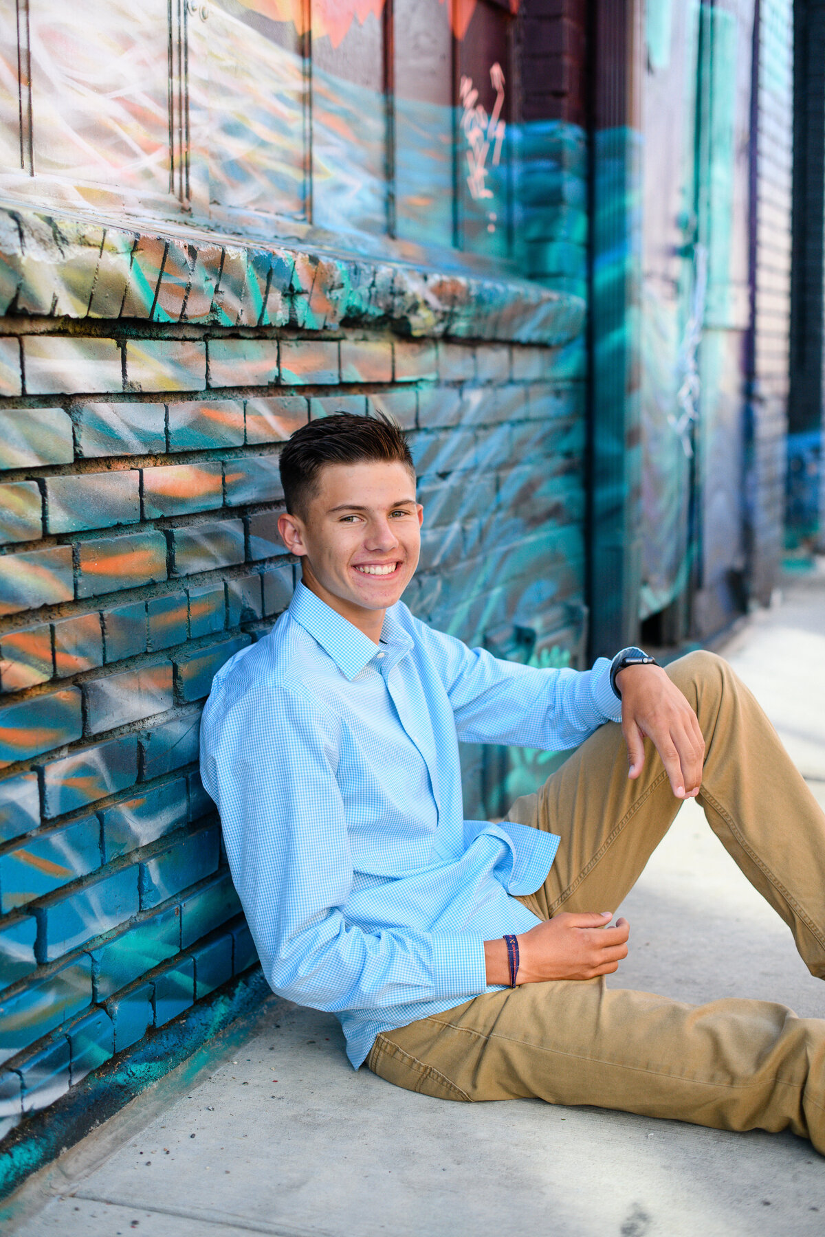 denver senior photographer captures senior guy photo ideas with young man sitting on the ground while wearing a blue button down and leaning against a wall mural in downtown denver with denver senior photographer