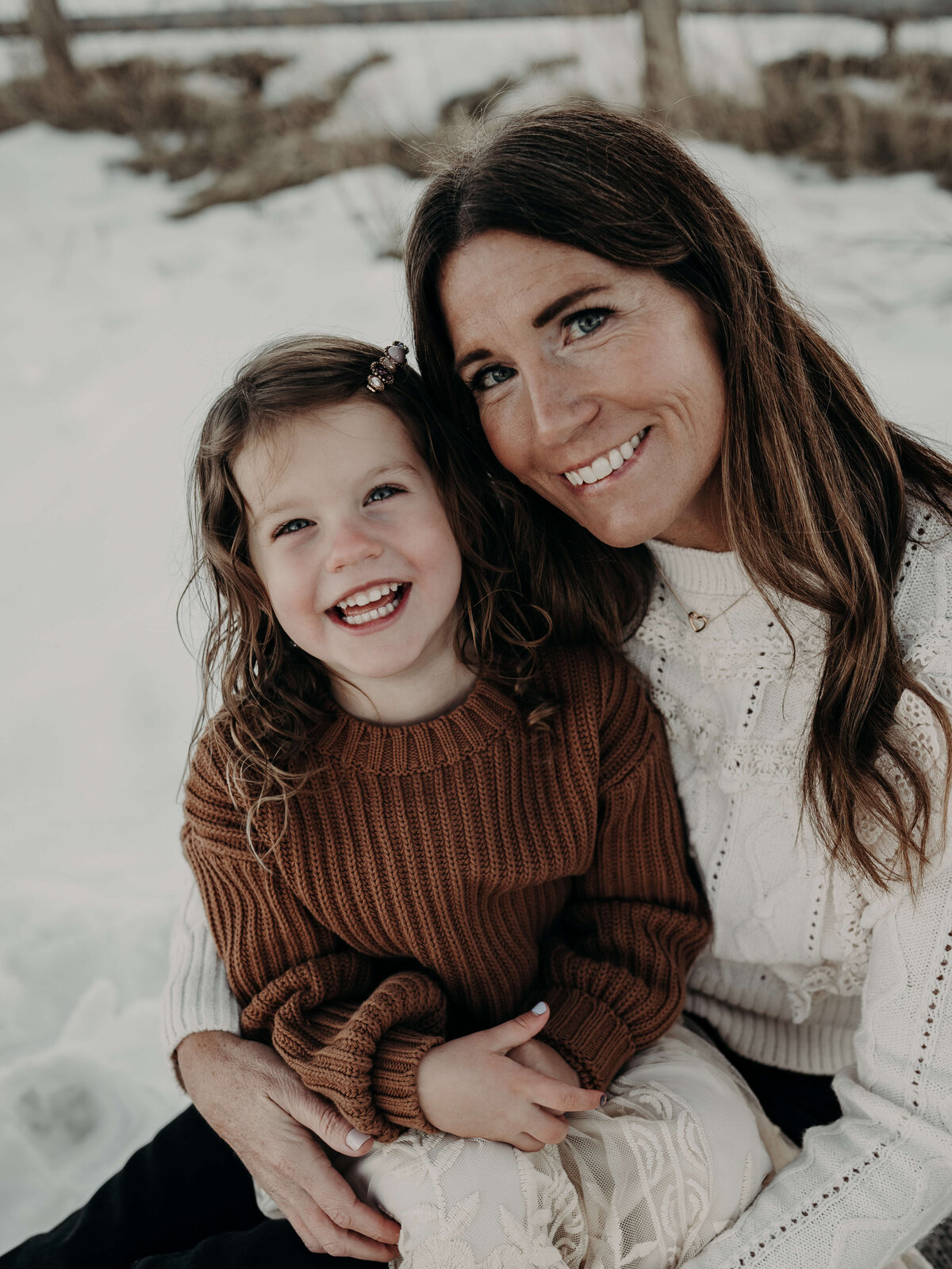 A beautiful mom holds her daughter that looks so much like her on her lap during a snowy outdoor family photo session in Denver, Colorado.