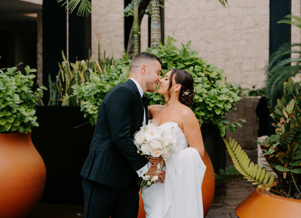 Bride and groom share a romantic kiss surrounded by lush greenery and tropical plants on their wedding day.