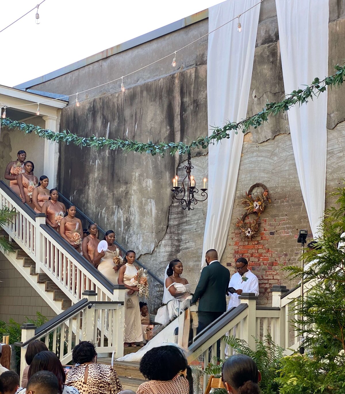 A bride and groom stand at the altar during an outdoor wedding ceremony. Bridesmaids in matching dresses line a staircase. Guests are seated in the foreground. The setting features draped fabric, greenery, and a brick wall.