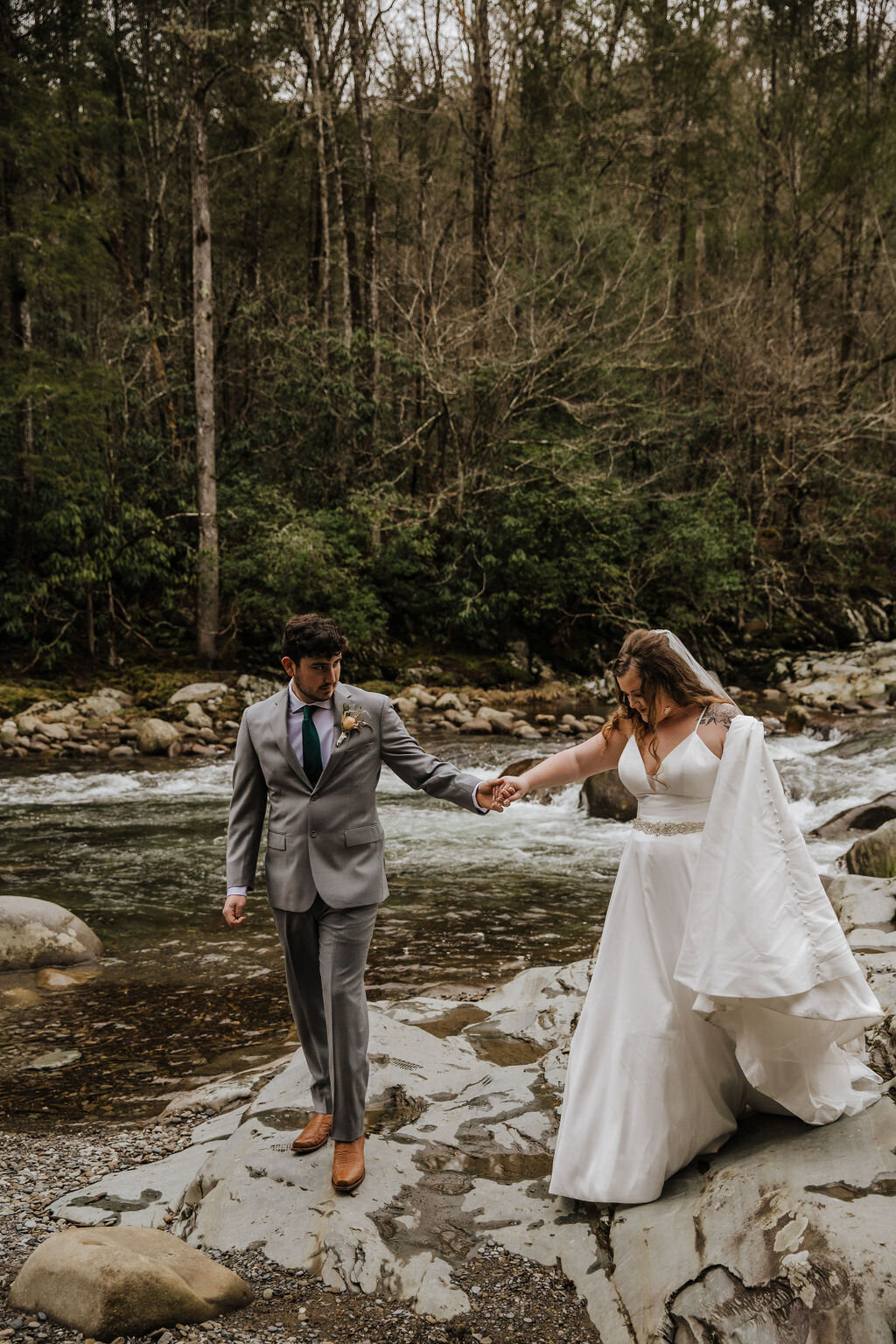 Bride and groom walking hand in hand across river rocks at Greenbrier after eloping to Gatlinburg, surrounded by forest and flowing water.
