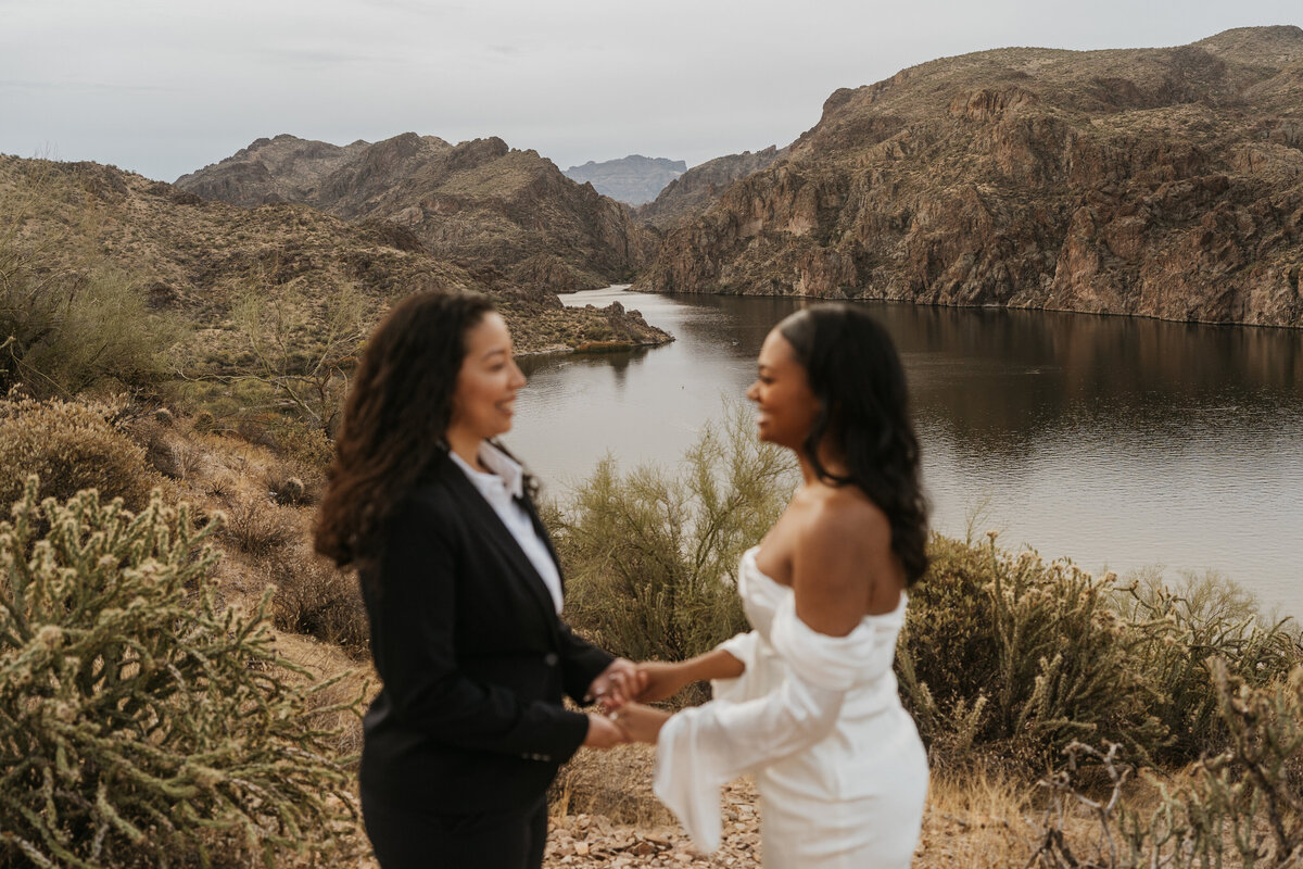 a newlywed couple holding hands while standing on a mountain overlooking a lake 