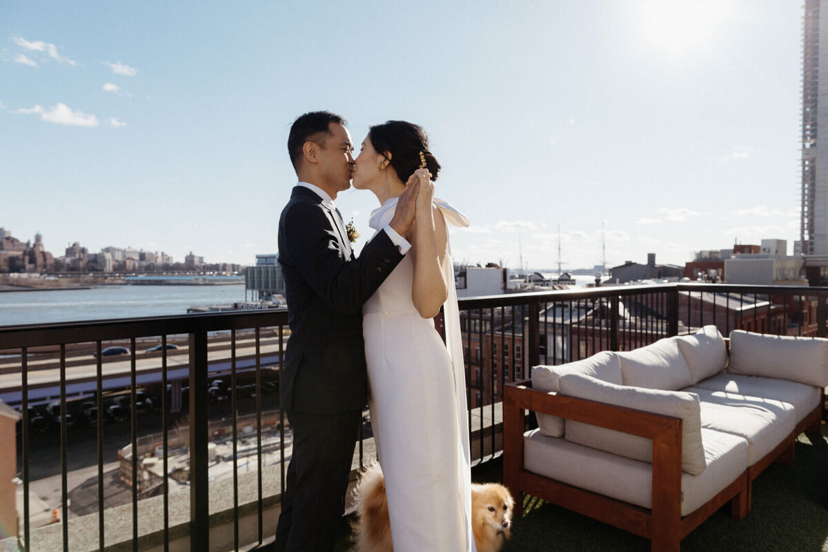 The bride and the groom hold hands while kissing, as they stand with their dog on a terrace overlooking Brooklyn.