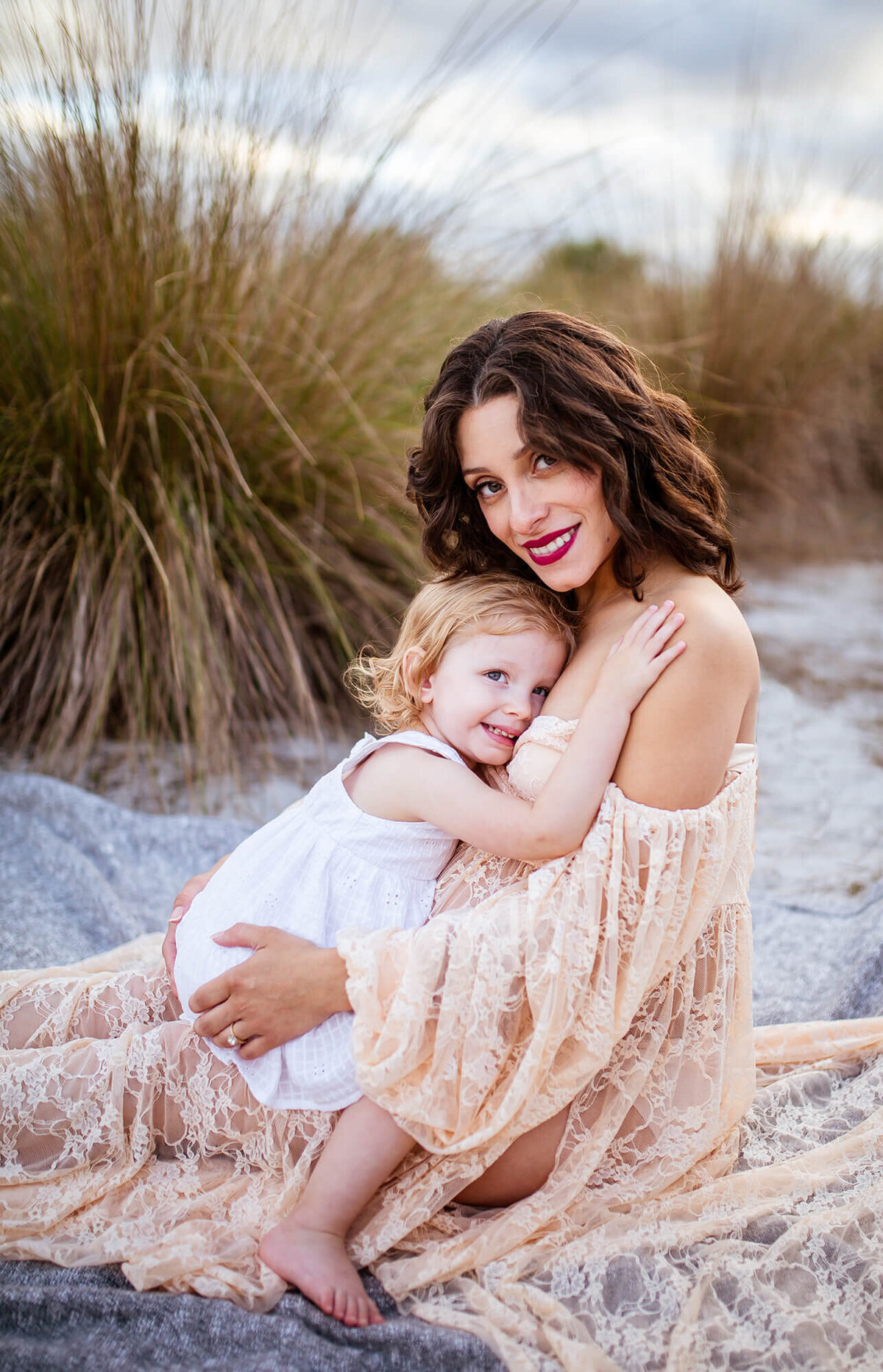 Woman sitting in tall grass cuddling small child on her lap. Captured by Sarah Wyndham Photography