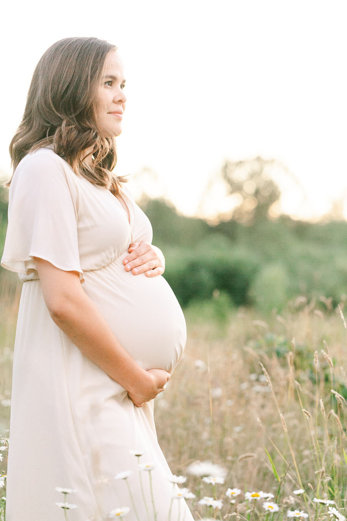 Profile image of a woman in cream dress holding  her baby bump and looking off into the sunset. She is standing in a field of golden grasses with some white wildflowers in the foreground.