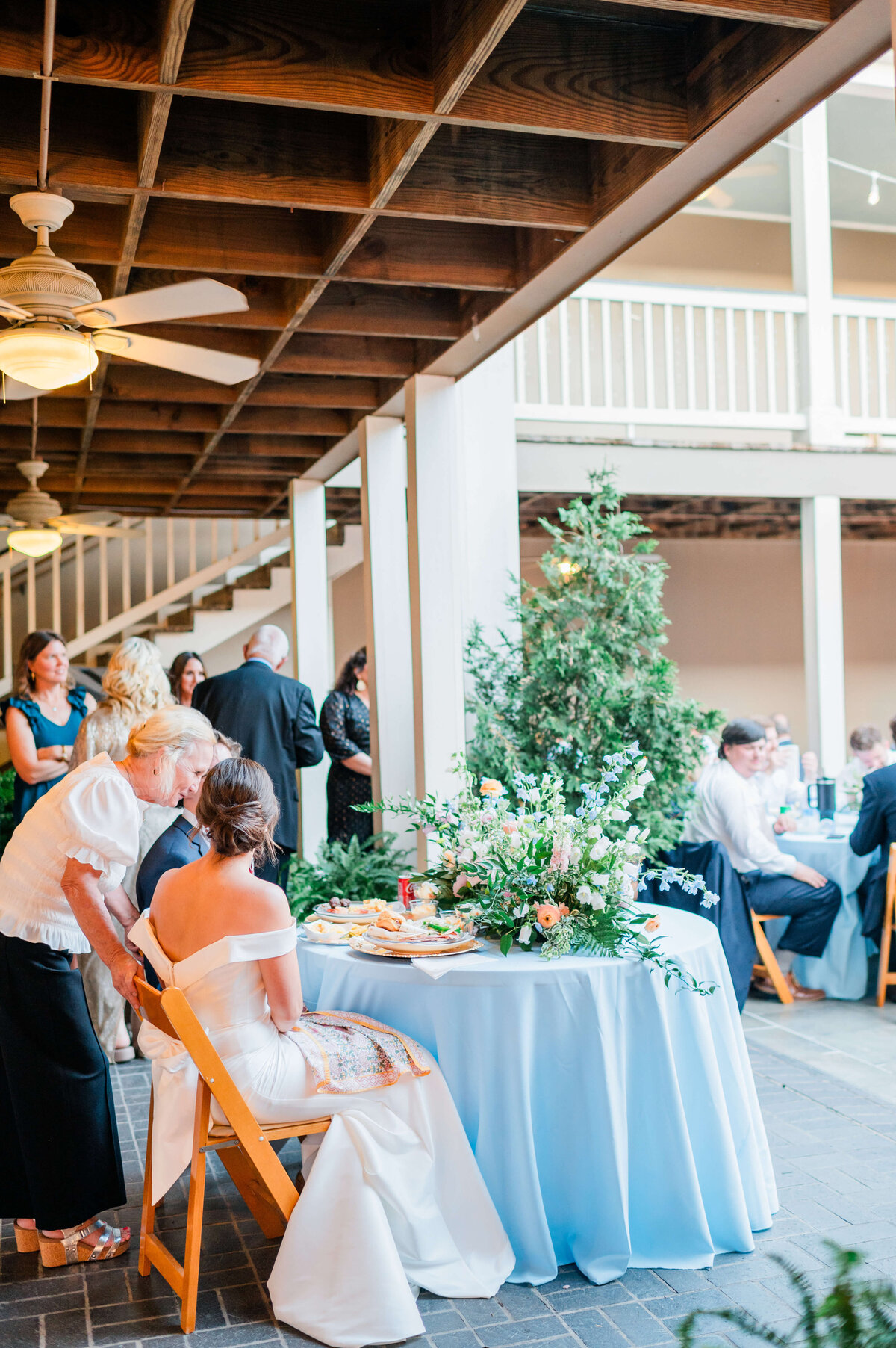 A bride in a white dress sits at a round table with a blue tablecloth and floral centerpiece while an older woman leans in to speak to her at a wedding reception with guests mingling in the background.
