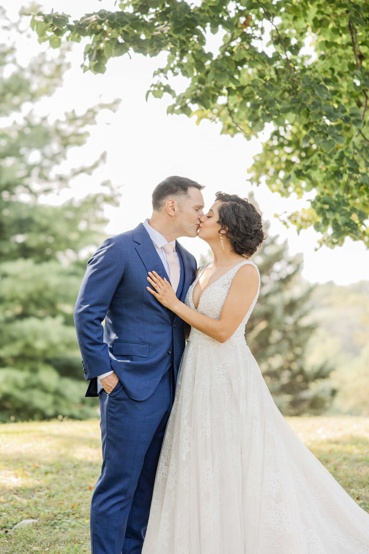 A couple in wedding attire kissing under a tree at an Iowa park, with the man in a blue suit and the woman in a white gown.