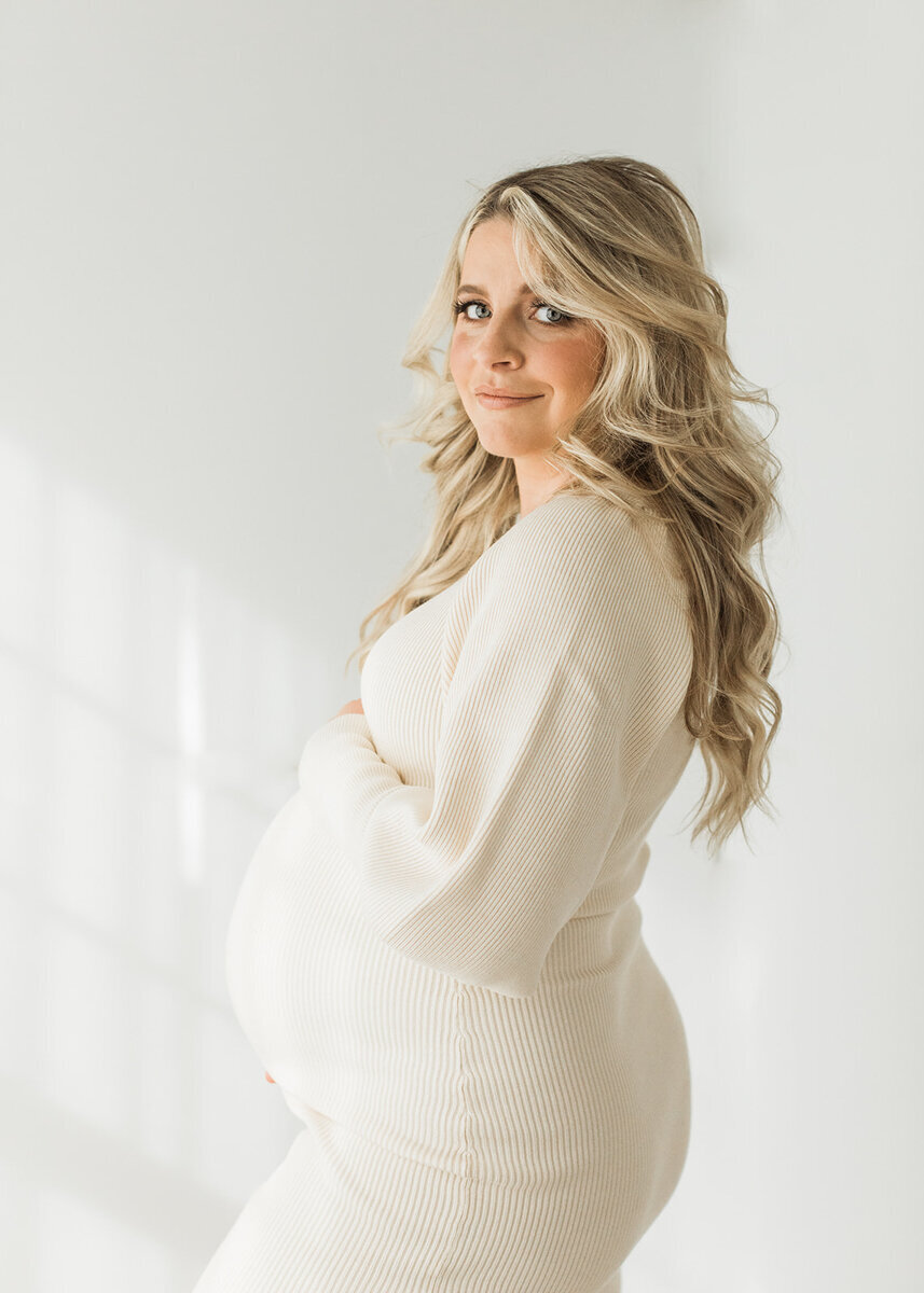 mom with long blonde hair stands in sweater dress showing off her pregnant bump