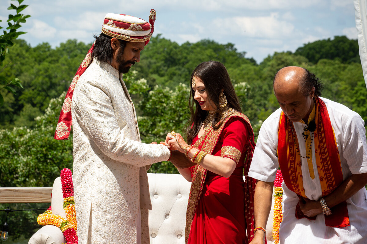 A bride and groom in traditional Indian attire participate in a wedding ceremony outdoors in Illinois, holding hands while a priest observes.