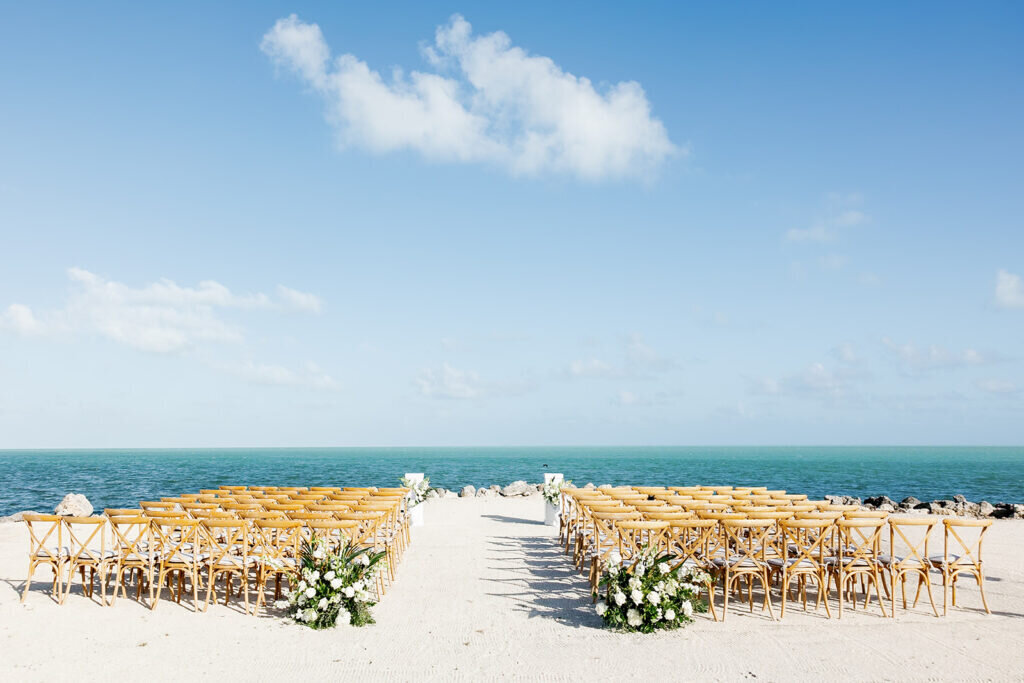 A breathtaking wedding ceremony setup on the beach, featuring wooden chairs, lush floral arrangements, and an oceanfront view. Destination wedding photography by Claudia Amalia Photography.