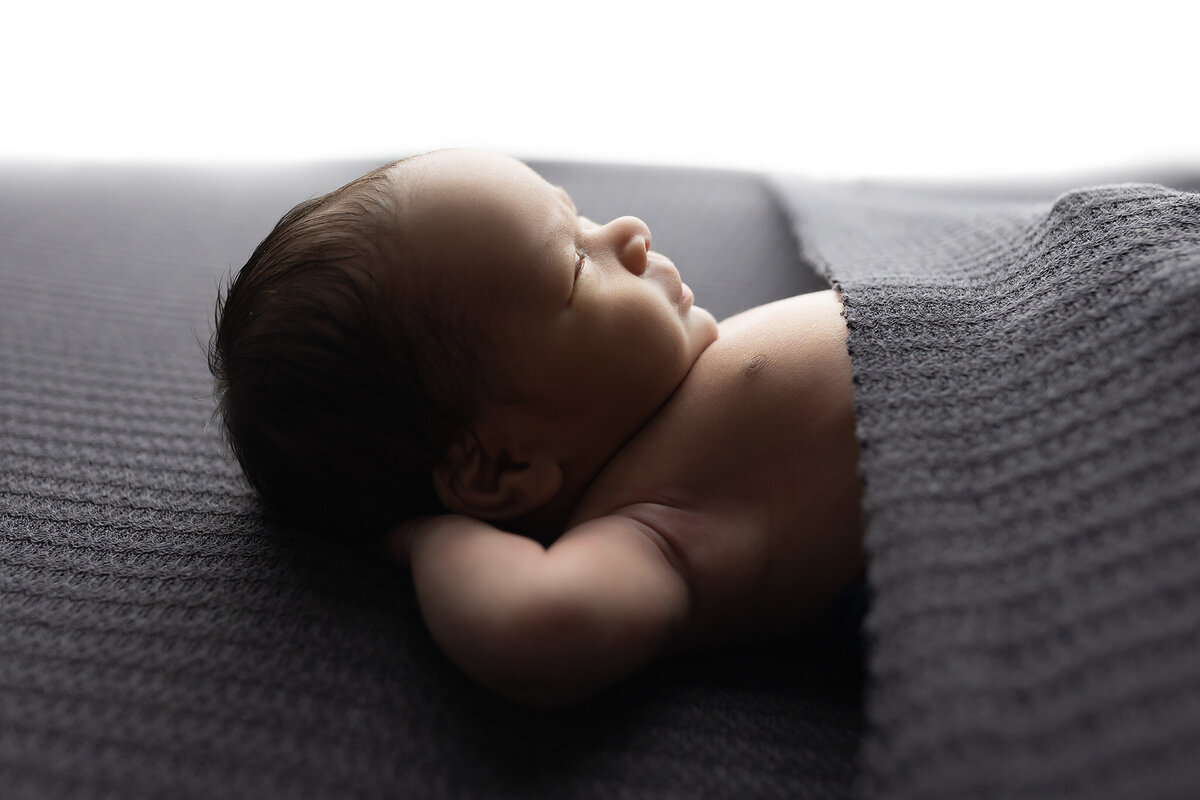 Baby lying on back under cozy gray knit with head turned — professional newborn photography.