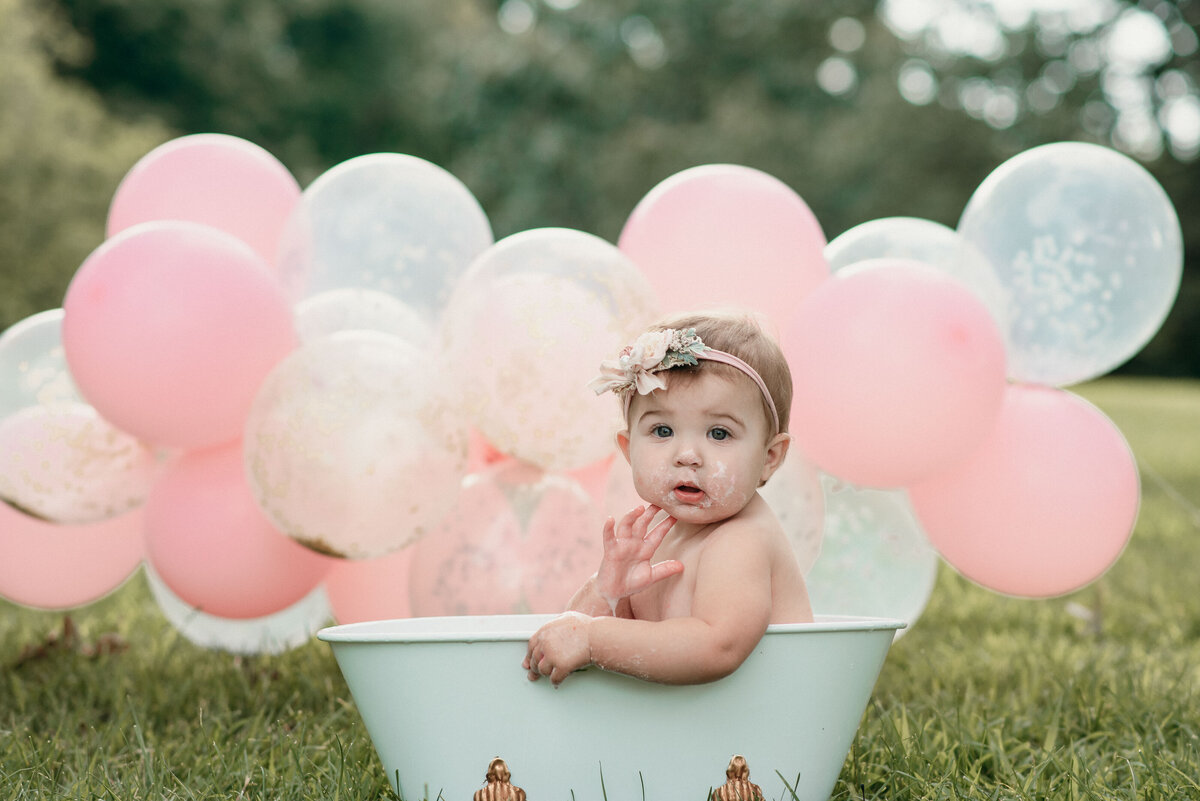 One year old baby girl outside sitting in mini bathtub wearing headband with pink and white balloon garland behind her