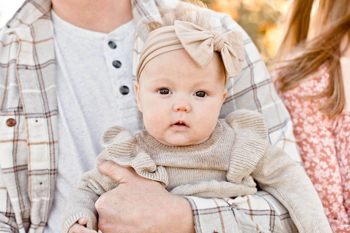 4 month old baby girl in beige sweater dress and beige bow looking at the camera. Dad is in a beige plaid shirt and is holding baby against his chest facing out with one arm under her arms. This image is a close-up of baby and dad's face is not in the picture.
