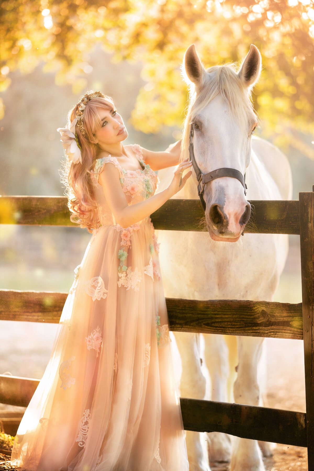 Asian woman with long blonde hair wearing a nude colored gown from Chotronette.  She is standing with a white Shire horse and petting her.  The horse is on the other side of a wooden fence.  There are golden leaves on the trees in the background and lots of sunlight.
