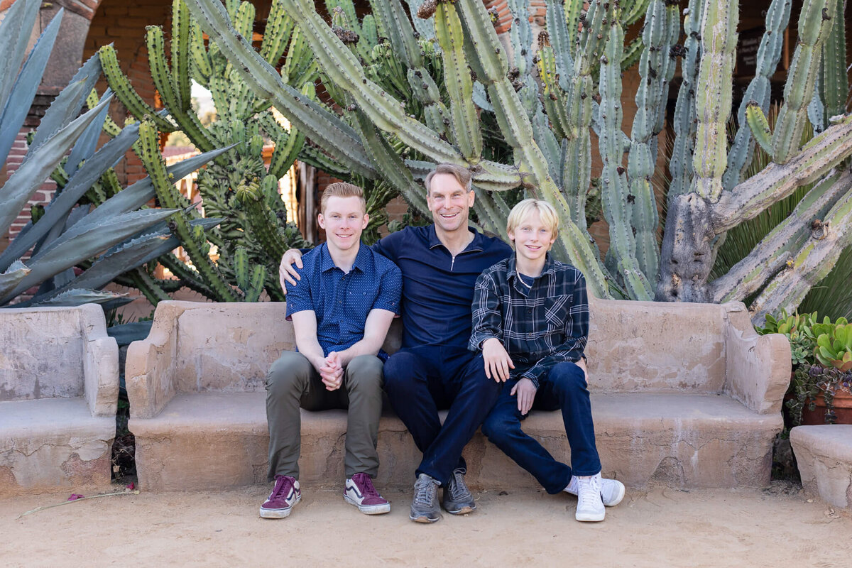 A father and his two teenage sons sitting on a clay bench with large cactus plants behind them, wearing shades of blue.