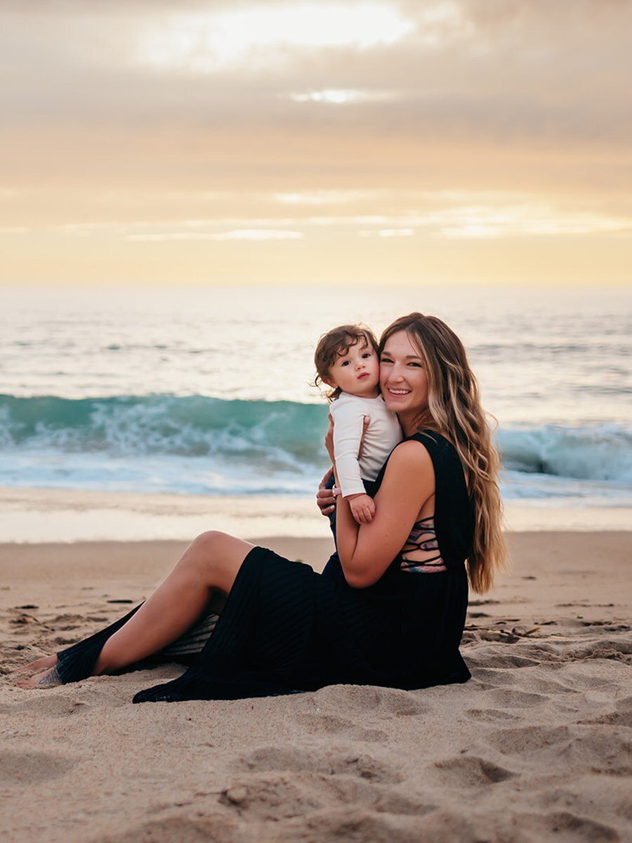 Smiling mother and baby sitting by the ocean at sunset — view family photo gallery