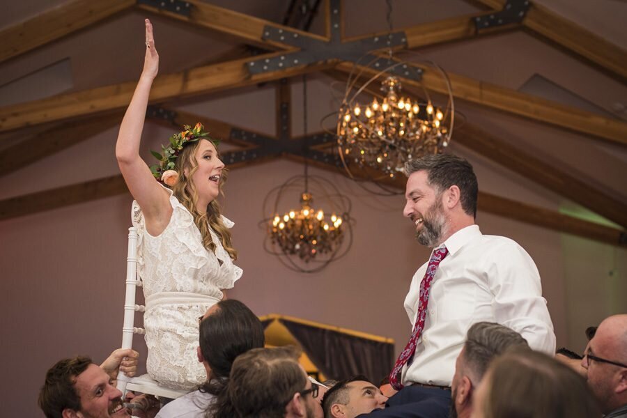 A bride and groom are lifted into the air by their wedding guests as they celebrate, and the bride throws her hands into the air.
