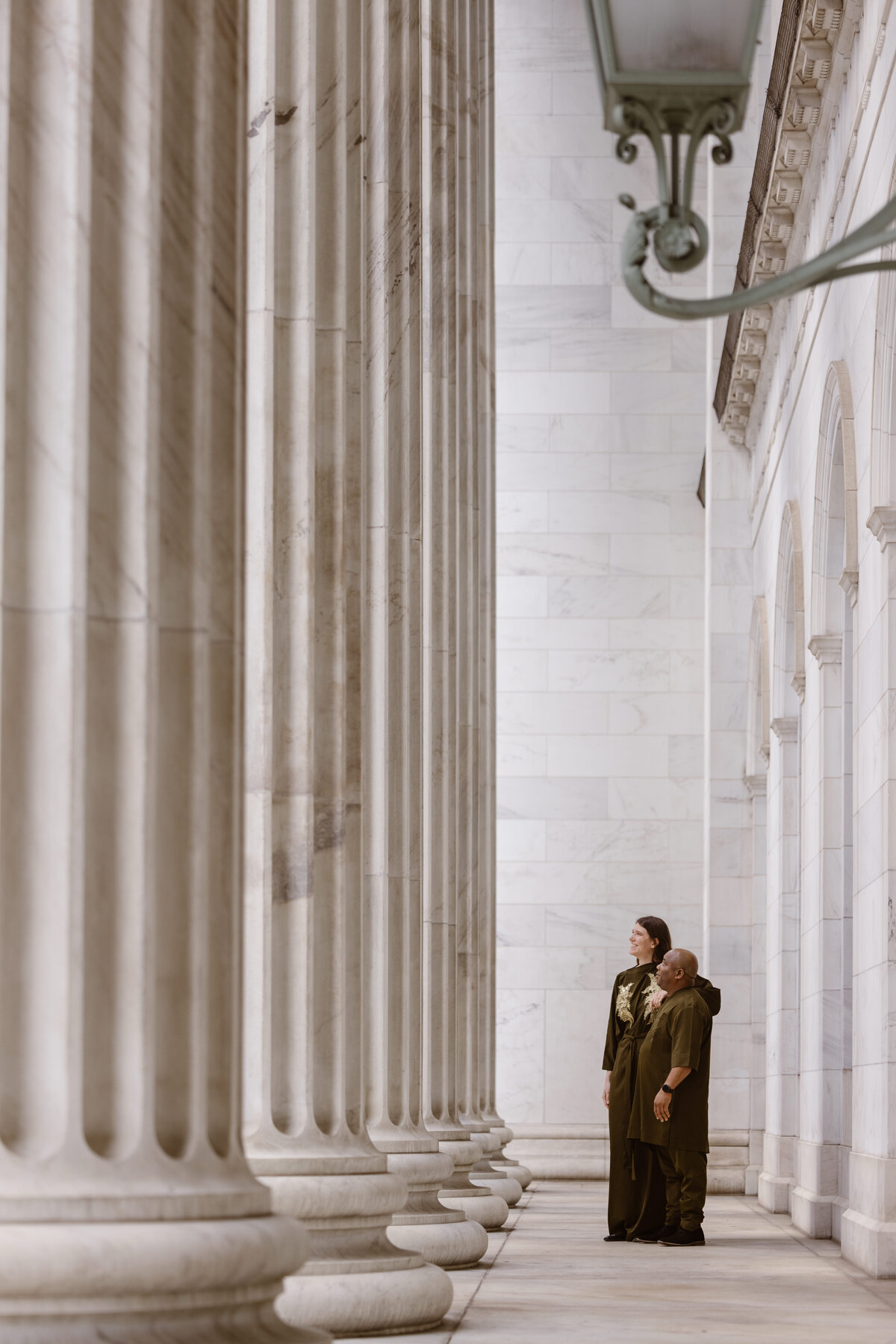 A couple stands closely together in a marble courthouse corridor lined with tall columns, looking upward with admiration.