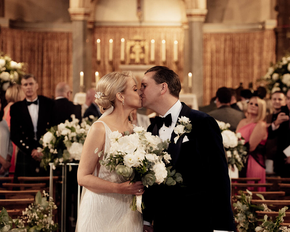 Bride and groom kiss in a cathedral after marrying. The bride holds her bouquet.