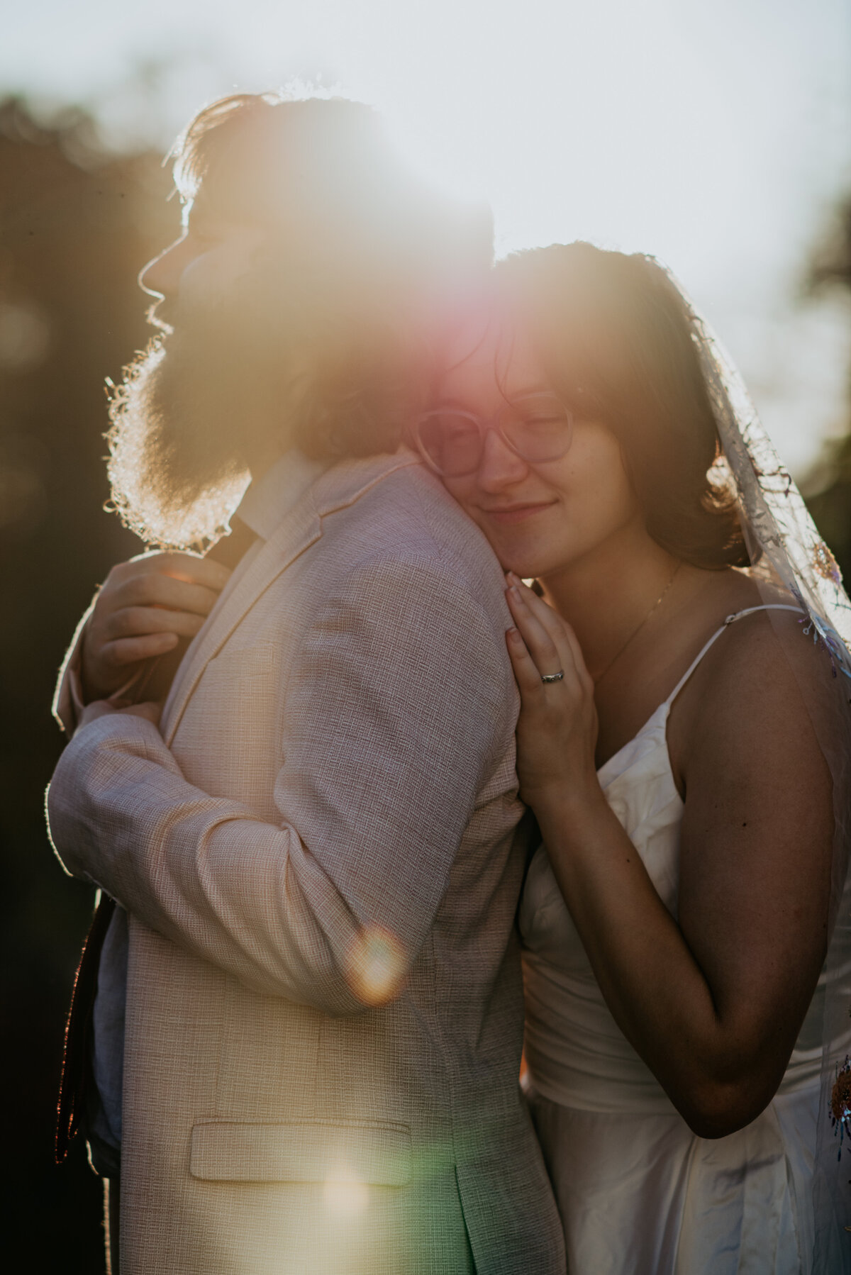 The bride leans against the groom's back as he adjusts his tie, the sun setting behind them and casting a flare.