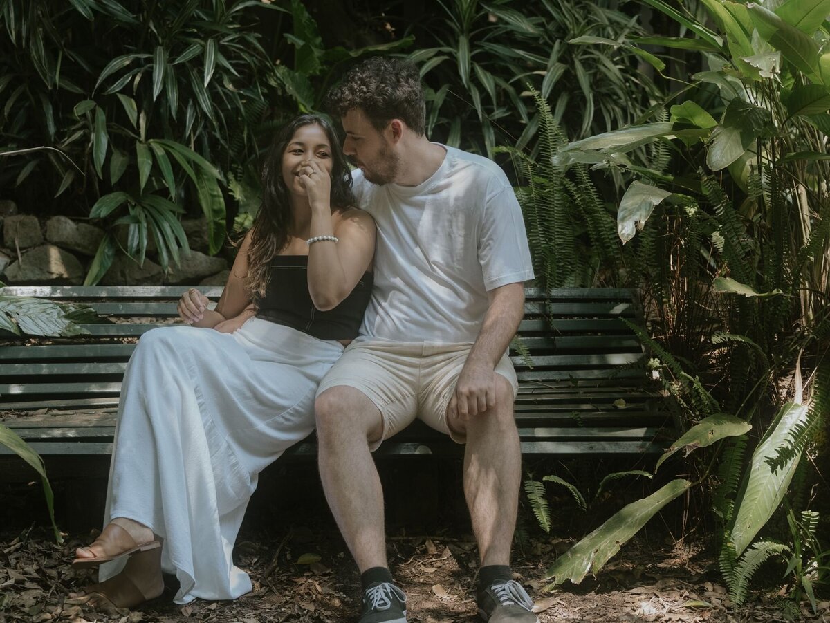 Engaged couple sitting on a park bench at Wendy’s Secret Garden in Sydney, sharing a candid romantic moment.