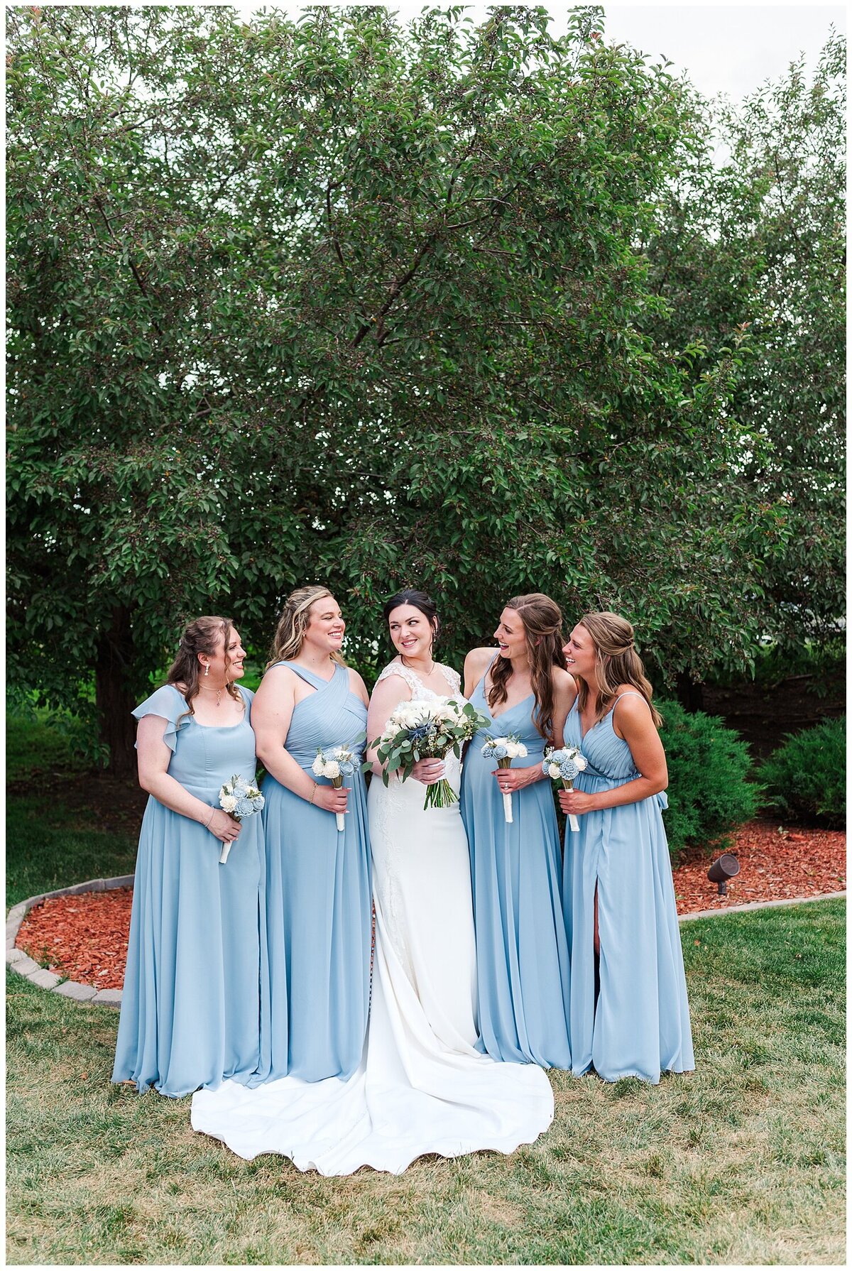 Four bridemaids in dusty blue dresses  stand next to a bride and exchange glances at each other