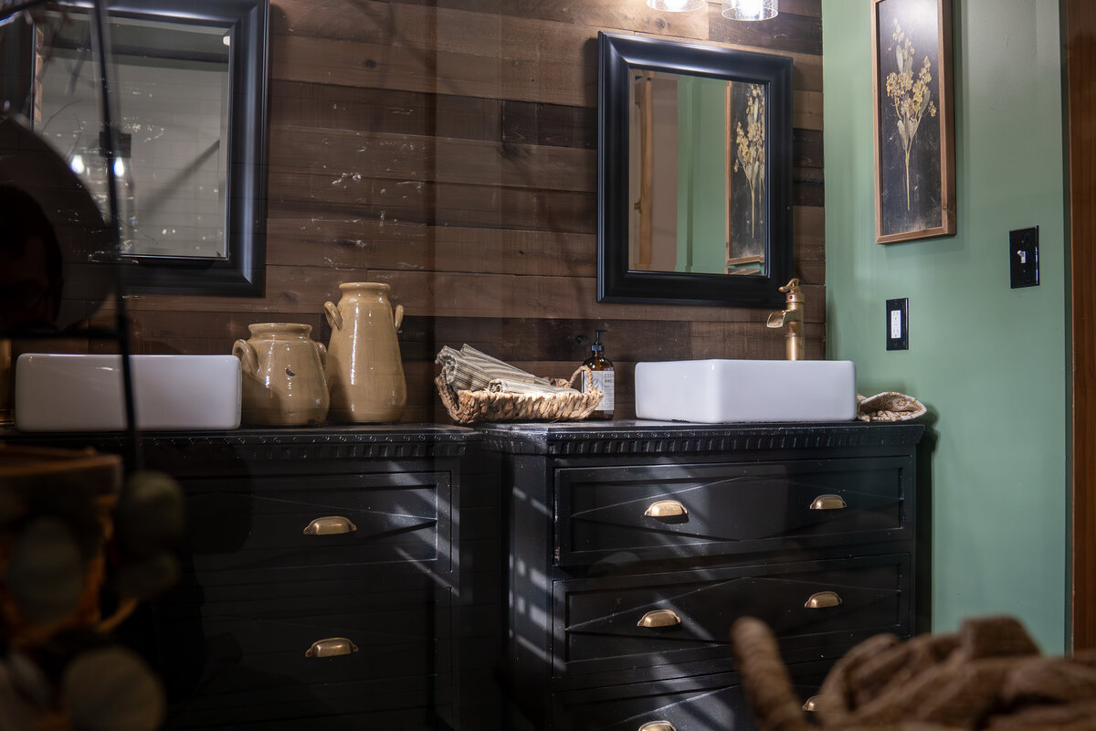 Rustic bathroom with wooden slat wall behind the black trimmed mirrors and black cabinet bureaus turned into bathroom vanities with white vessel sinks