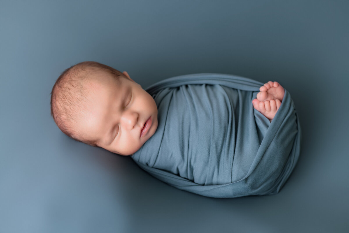 Newborn Photographer, a baby lays on blue sheets waddled in a blue wrap