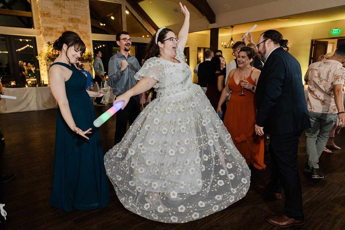 A bride cheering and dancing with her wedding guests during her wedding reception at The Laurel in Grapevine, Texas. The bride is wearing a reception dress that is long, flowing, and white with a floral headband and is holding a foam light stick. Her guests stand around her and cheer her on.