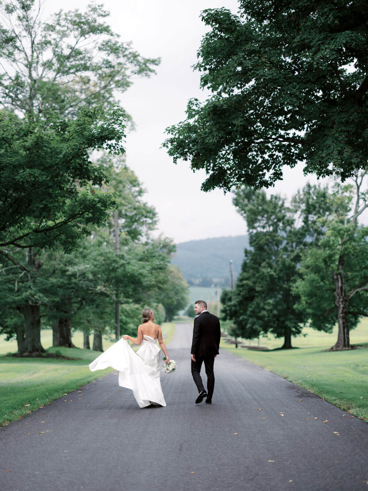 Back view of the bride and groom walking on a picturesque road at Lion Rock Farm, Sharon, CT.  Image by Jenny Fu Studio