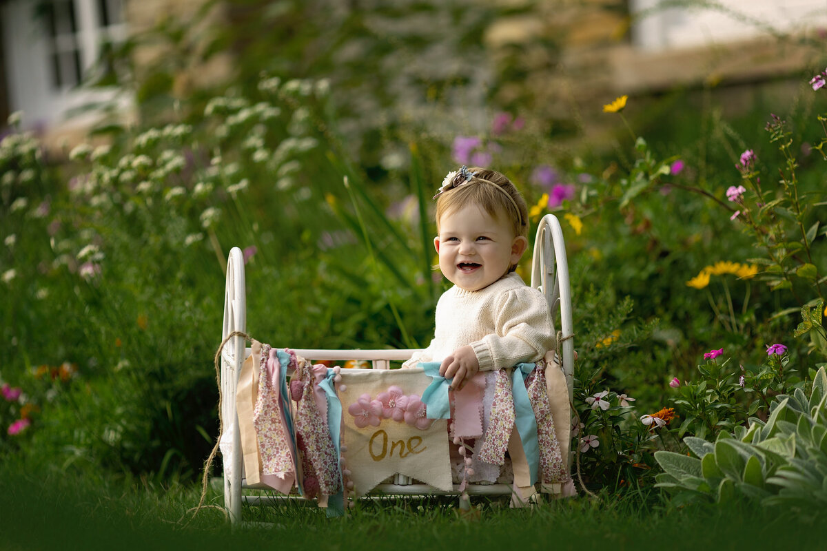 A toddler girl sits in a crib in a garden decorated for her first birthday
