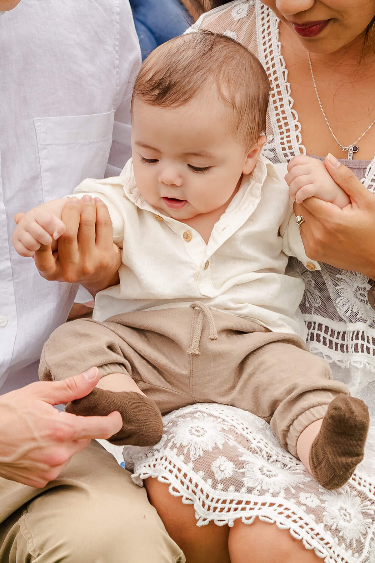 A baby wearing neutrals sits on his mom's lap while his dad tickles his foot.