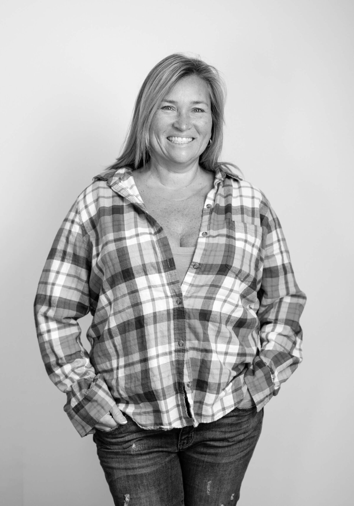 Before shot of a woman in a plaid shirt, hands in pockets, smiling with a friendly demeanor.