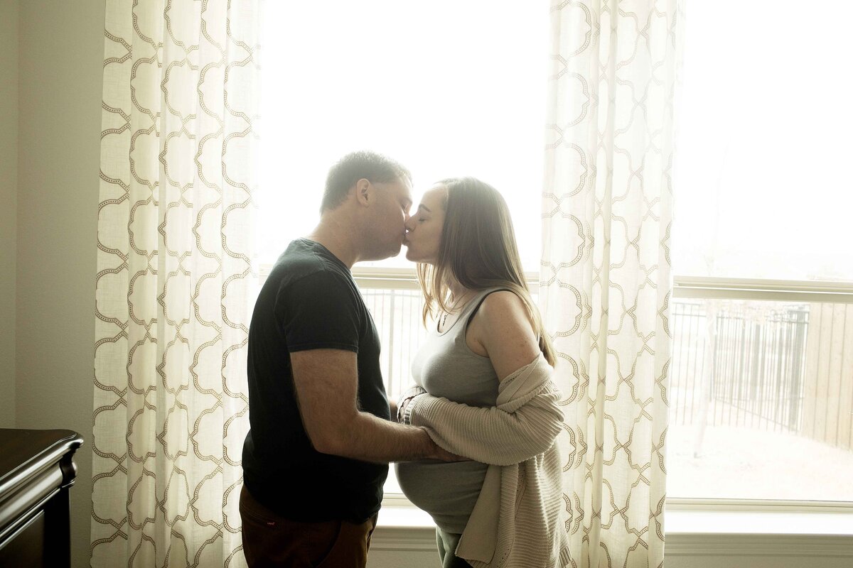 A heartwarming maternity shot of mom and dad standing together in front of a window, with soft sunlight casting a glowing halo around them, creating a quiet, intimate moment, captured by Chunky Monkey Photography.