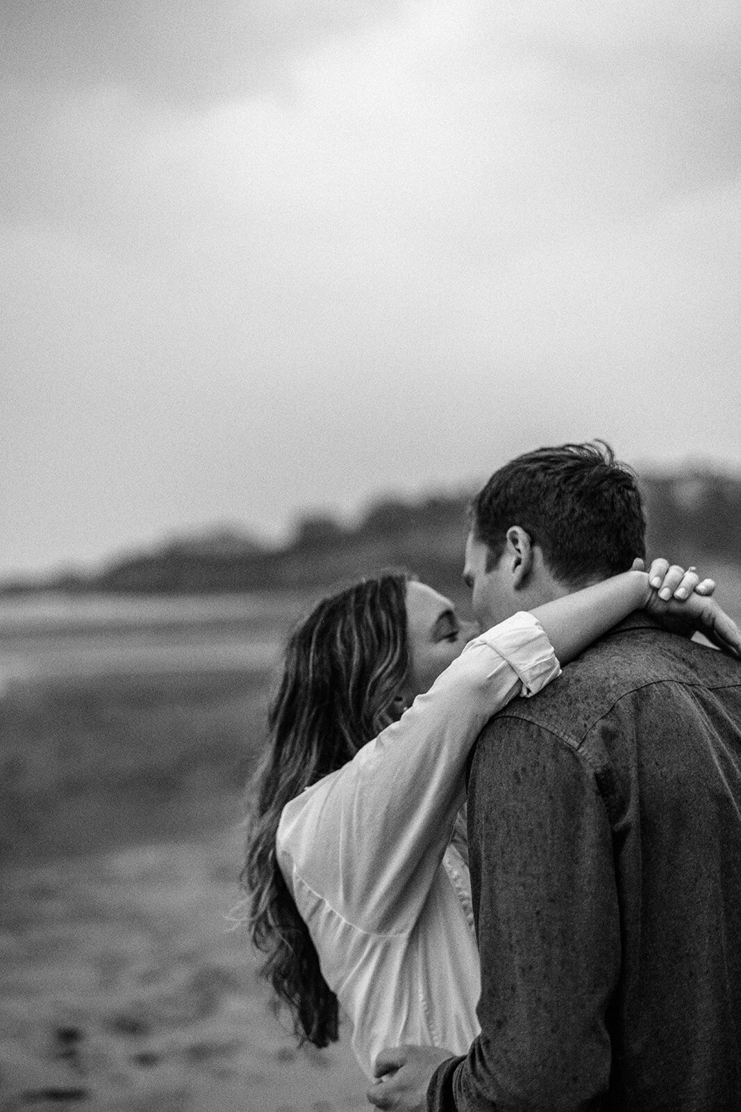 A woman with her arms around a man's neck while the kiss at the beach.