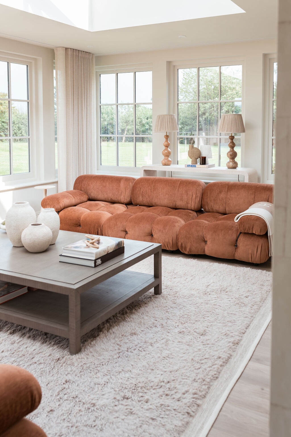 A cozy living room with large windows. The room features two rust-colored, plush sofas, a light grey coffee table with vases and books, and a soft, white rug covering the floor. Two lamps are on side tables flanking a large sofa.