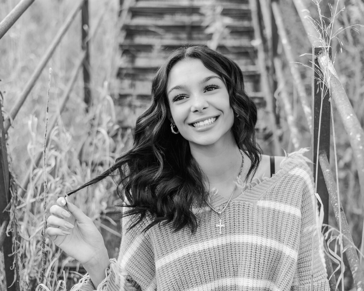 black and white photo of a female high school senior with long dark hair, sitting, looking away from the camera smiling while playing with her hair, wearing a striped shirt