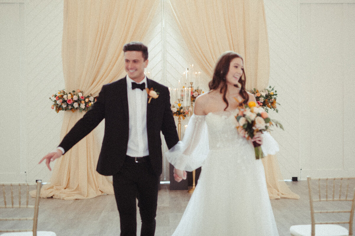 A bride and groom wearing a white wedding gown and black tuxedo walk down the aisle holding hands.