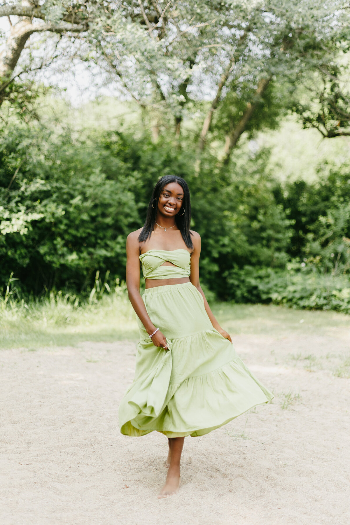 Senior girl in a matching green top and skirt set twirling her skit and walking in front of green trees